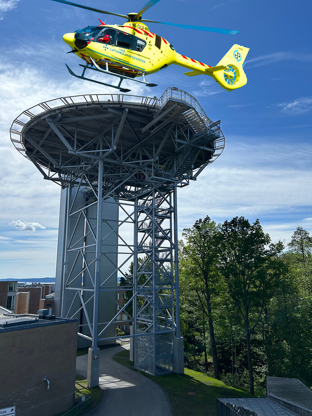 Heliport ved Rikshospitalet i Oslo sett fra siden med et gult ambulansehelikoper som holder på å lande på plattformen.