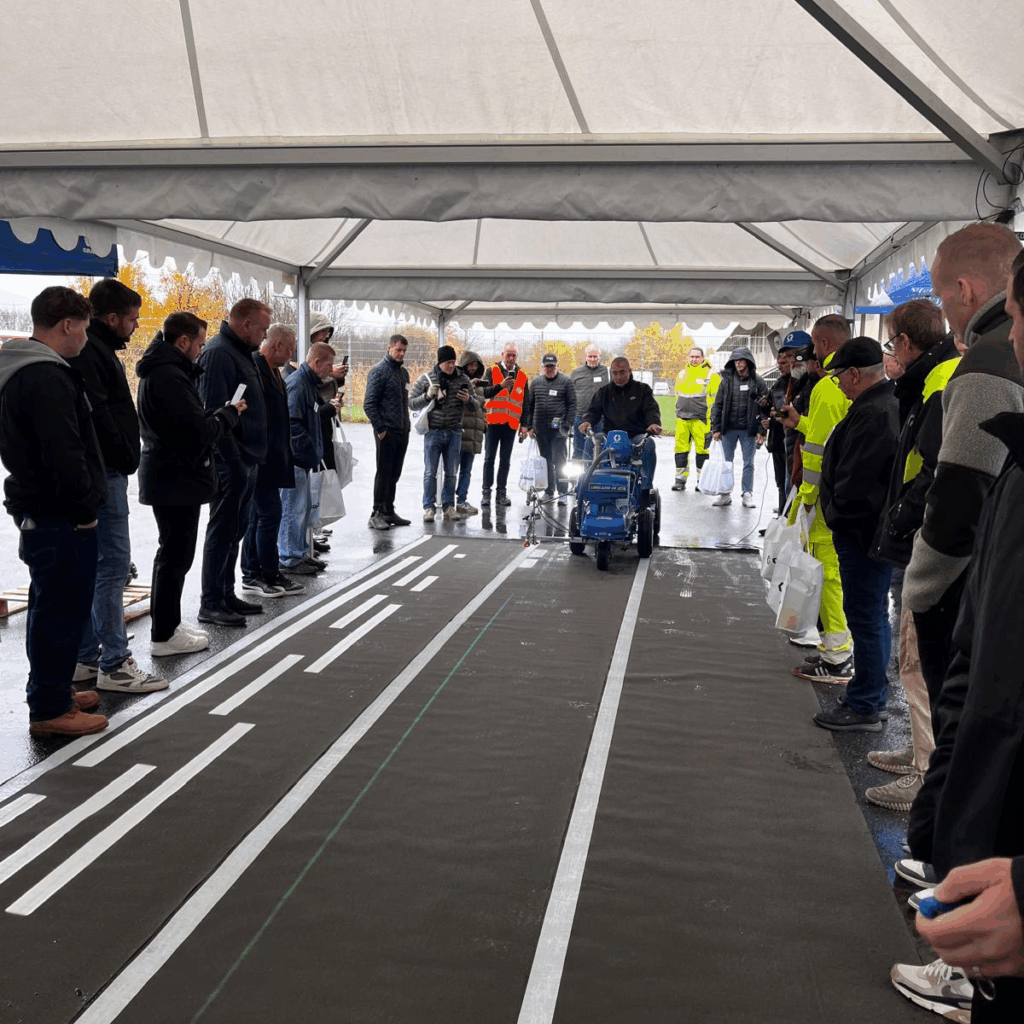 Mennesker på venstre og høyre siden av et langt teppe som ser ut som asfalt. I bakgrunn en mann på stripemaskin som kommer mot kameraet