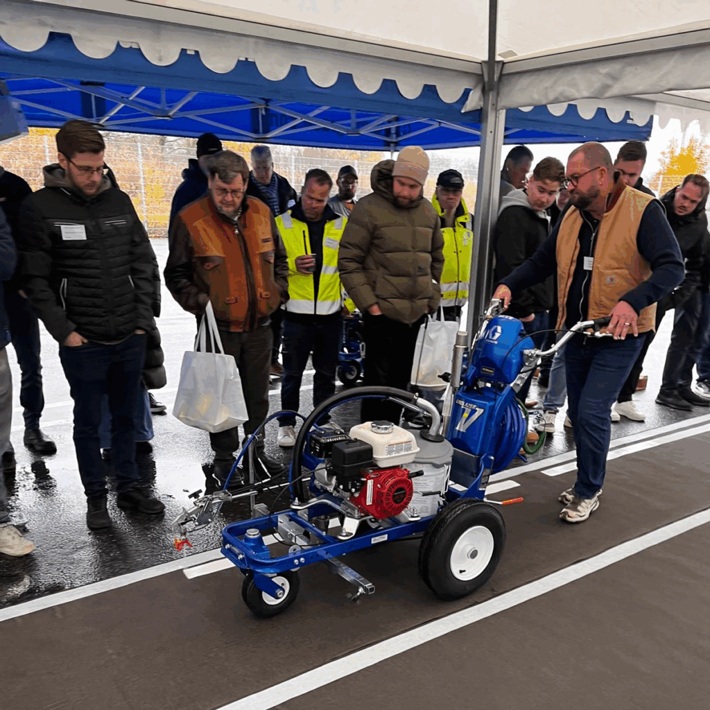 Mennesker i bakgrunn foran et langt teppe som ser ut som asfalt. I forgrunn en mann som skyver en stripemaskin