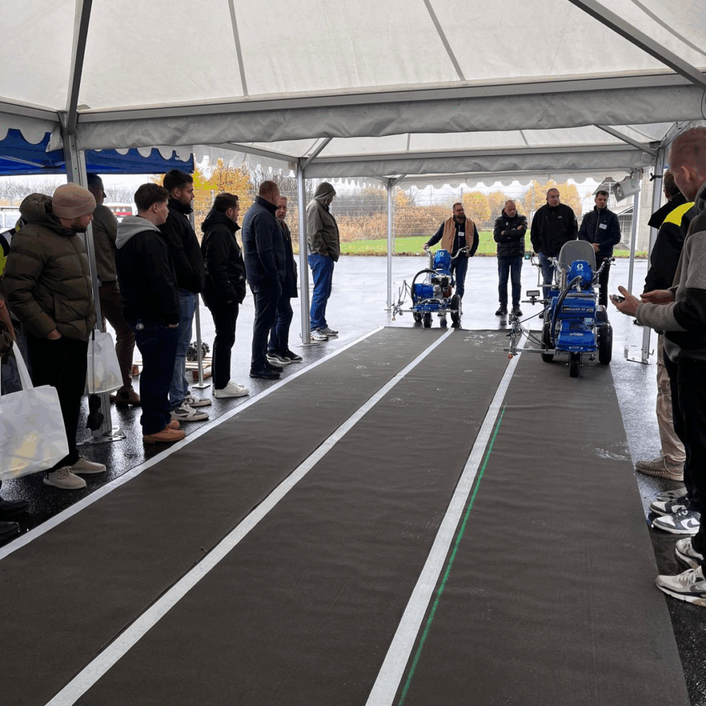 Mennesker på venstre og høyre siden av et langt teppe som ser ut som asfalt. I bakgrunn en mann på stripemaskin som kommer mot kameraet