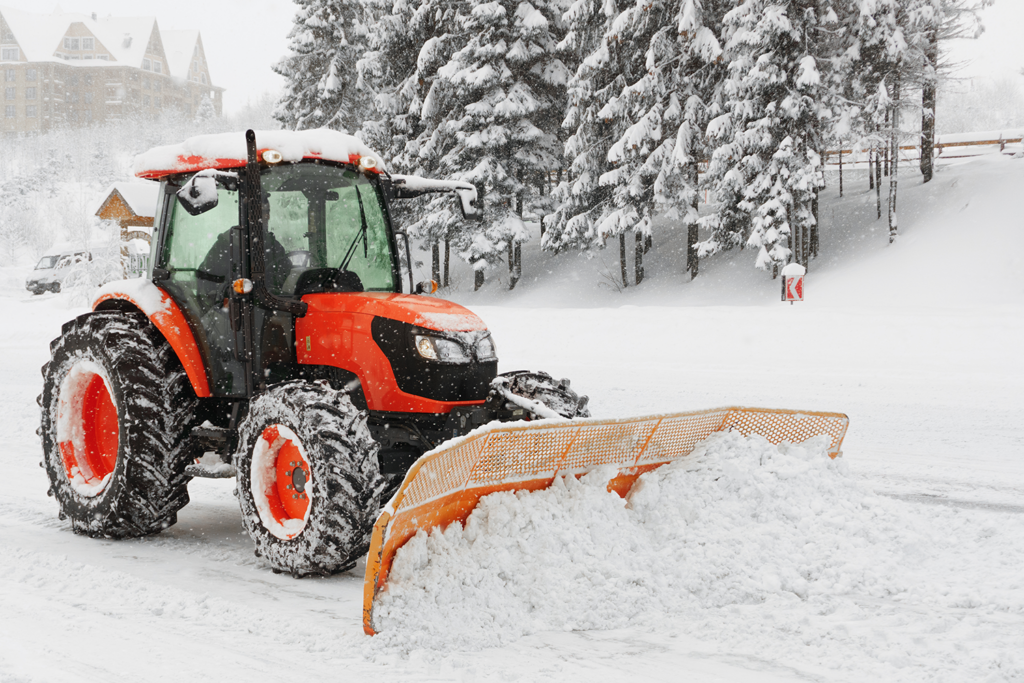 rød Kubota traktor med snøskuffe foran som måker snø. Trær i bakgrunn, alt er dekket med snø