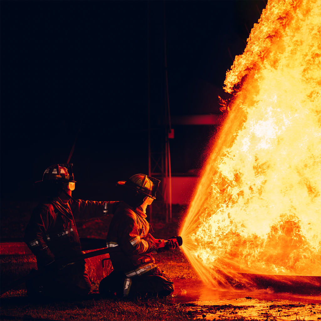 to brannmenn kneler på bakken og retter en brannslange mot brann. Bakgrunn er svart natt.