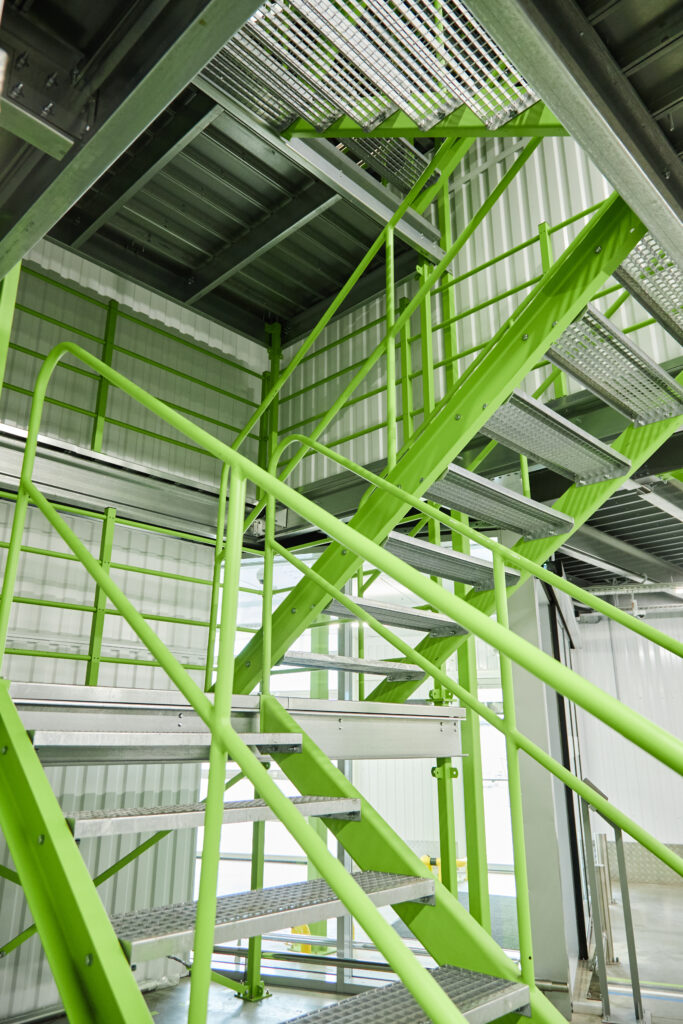 This image captures a modern industrial staircase within a building. The stark, geometric design features green metal railings and grated metal steps, creating a visually striking architectural element. The stairs lead to upper levels within the industrial space. Natural light filters in, adding to the clean and functional aesthetic. This image would be well suited for projects focusing on architecture, industrial design, construction, or urban environments, offering a fresh and contemporary visual.
