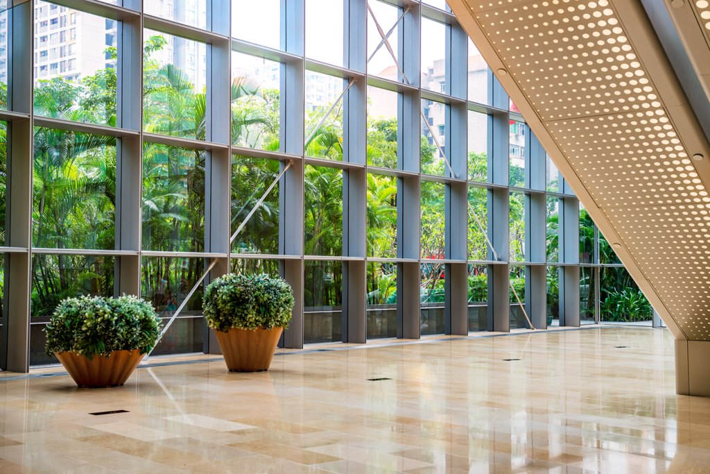 A naturally lit, inviting interior features abundant foliage visible through massive windows. Two green potted plants sit in the foreground on a reflective tan tile floor. The large windows show a dense garden with towering trees just outside the glass. The lobby's contemporary architecture is enhanced by an overhead lighting fixture with a distinct pattern.