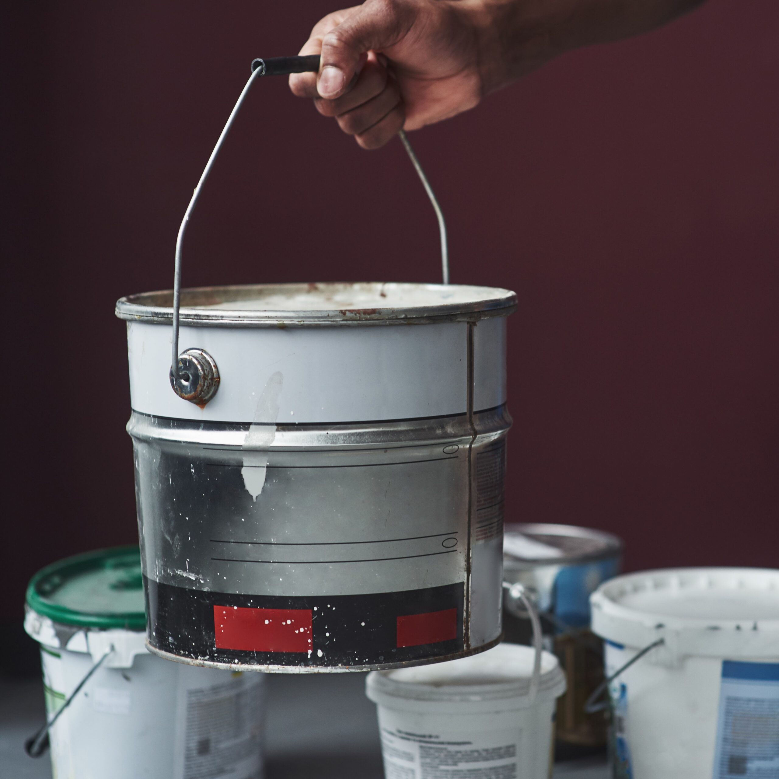 en hånd som holder et malingsspann i kameraet. Flere malingsspann på gulvet i bakgrunn