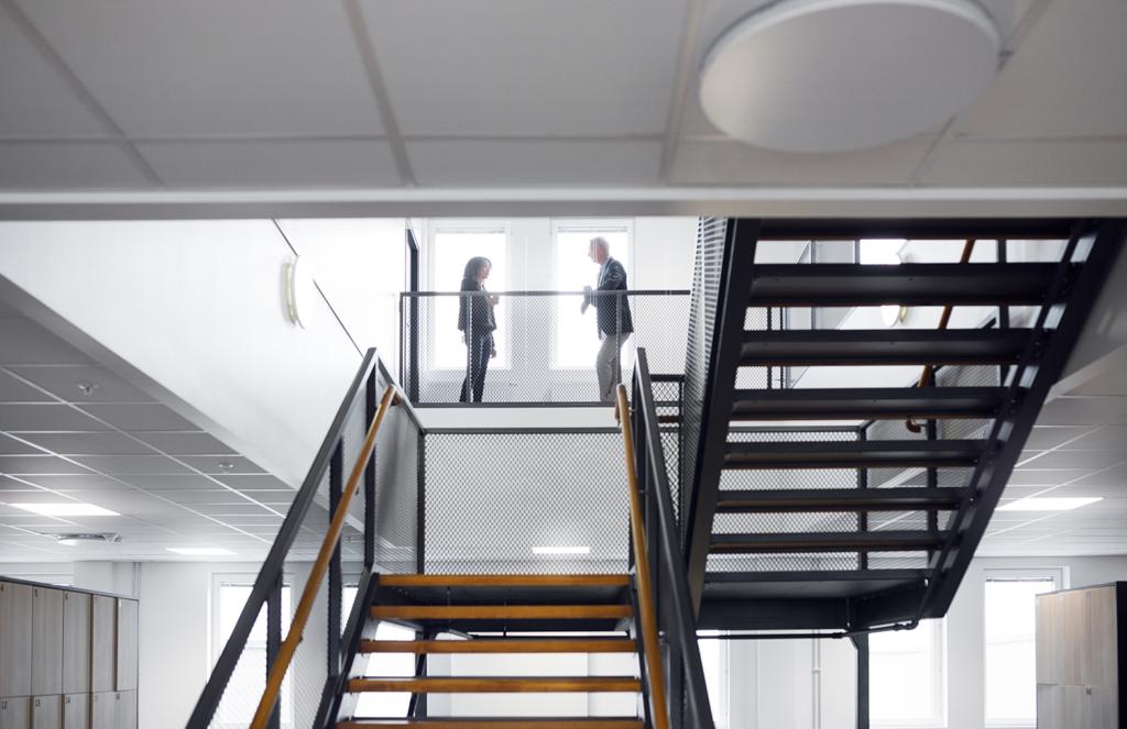 In a brightly lit office setting, a woman with dark hair and a man with light hair are seen on a balcony, engaged in a conversation. Both are dressed in business attire. The setting features industrial-style metal and wood stairs