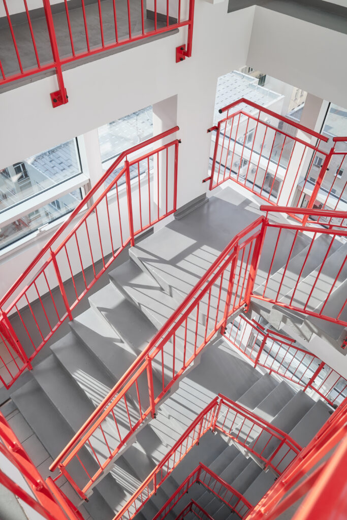 This striking architectural photograph captures a multi-level indoor staircase featuring vibrant red metal railings contrasted against smooth grey concrete steps and white walls. Shot from a high-angle perspective, the composition emphasizes geometric patterns and the vertical depth of the stairwell. Natural light streams in through large windows, casting sharp shadows of the railings onto the landing, creating a clean and industrial aesthetic.