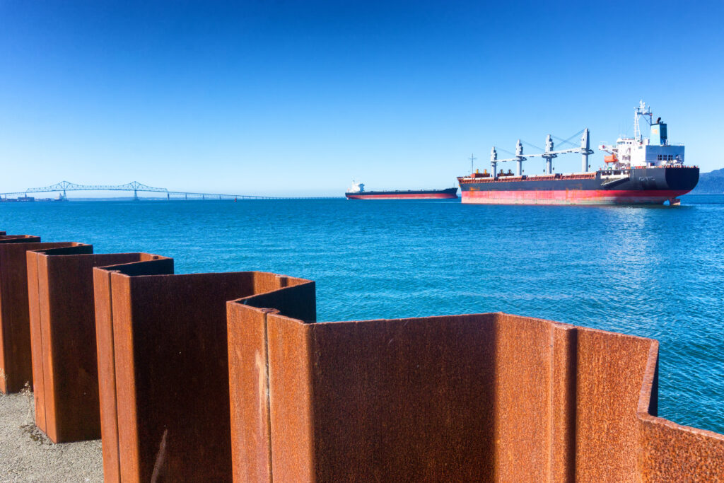 A picturesque seascape features a serene, azure sea under a clear blue sky. In the foreground, a series of rusted metal barriers lines the shore. Two massive cargo ships sail towards the horizon, one closer than the other.